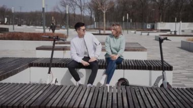 Bir adam ve bir kadın bankta oturmuş sohbet ediyorlar. Çiftin yanında elektrikli scooterları var..