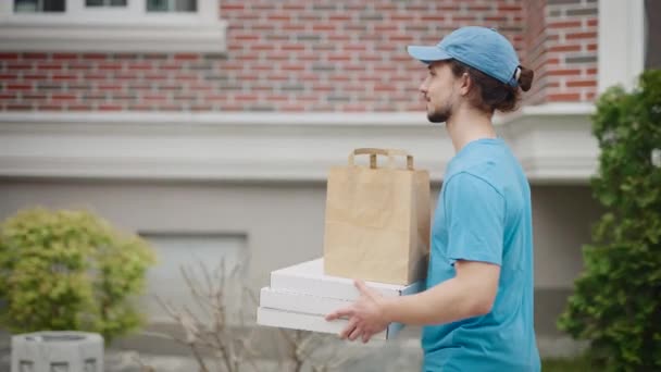 Un tânăr care livrează mâncare trece printr-un cartier modern și plăcut. Un barbat cu capac si tricou livreaza pizza si alimente — Videoclip de stoc