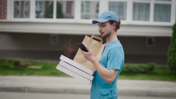 Um jovem entregador na rua com uma ordem. Um homem procura o endereço desejado usando seu smartphone. — Vídeo de Stock
