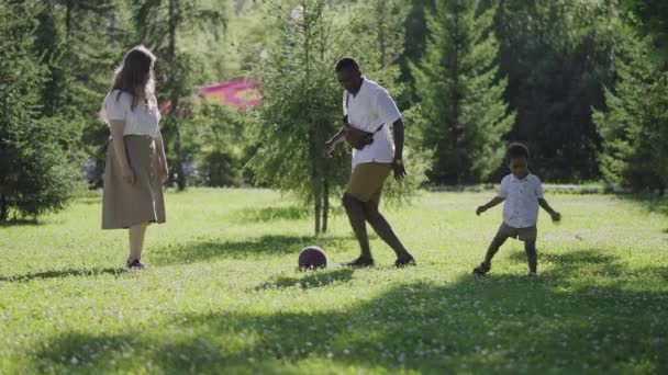 Familia multiétnica en el parque. Mamá rubia y afroamericana marido jugar al fútbol con su hijo. — Vídeos de Stock