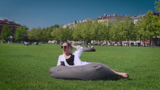 Bella bionda nel parco cittadino. Una donna tiene uno smartphone tra le mani e giace su un cuscino. — Video Stock