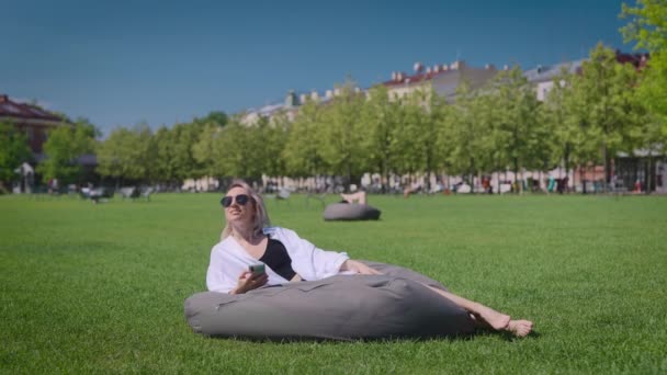 Гарна блондинка в міському парку. Жінка тримає в руках смартфон і лежить на подушці . — стокове відео