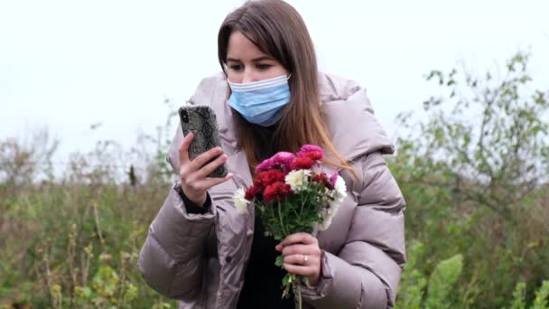 Fille dans un masque médical bleu avec des fleurs dans ses mains parle sur un smartphone. — Video