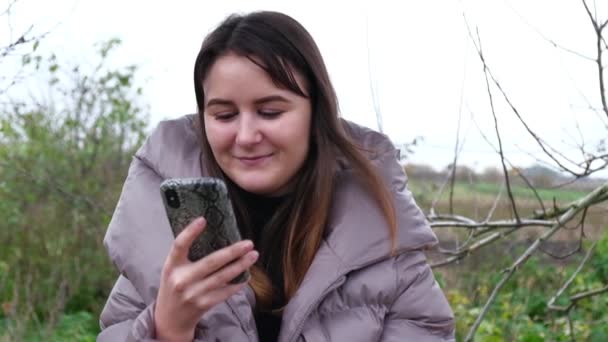 Menina vestindo um casaco quente sentado no parque com um smartphone — Vídeo de Stock