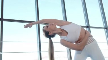 Beautiful sports girl in tight sportswear performing exercises on a mat in a bright studio with large French windows.