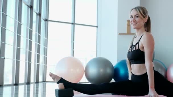 Belle fille dans un uniforme de sport étirant ses jambes sur un tapis dans la salle de gym. — Video