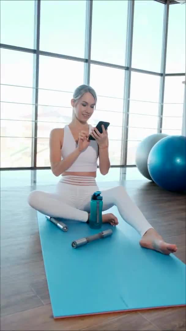 Glad tjej i sportkläder sittandes på yogamattan medan du använder smartphone — Stockvideo