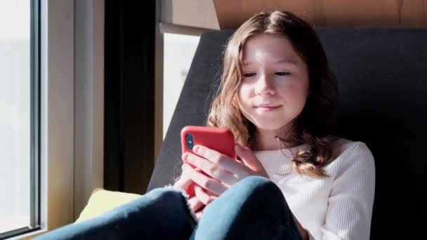 Belle petite / jeune fille joyeuse assise près de la fenêtre avec le téléphone. — Video