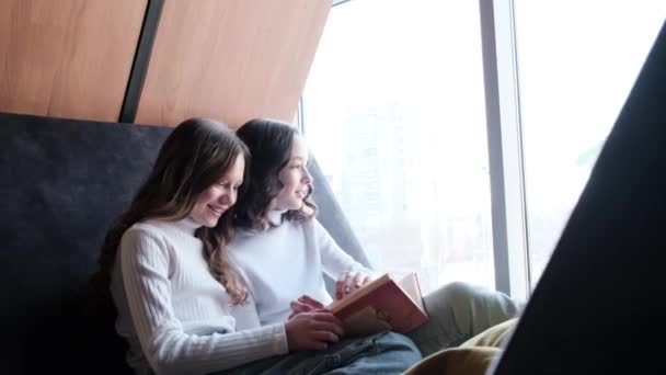 Two girlfriends sitting on a sofa by the window flipping through a book. — Stock Video