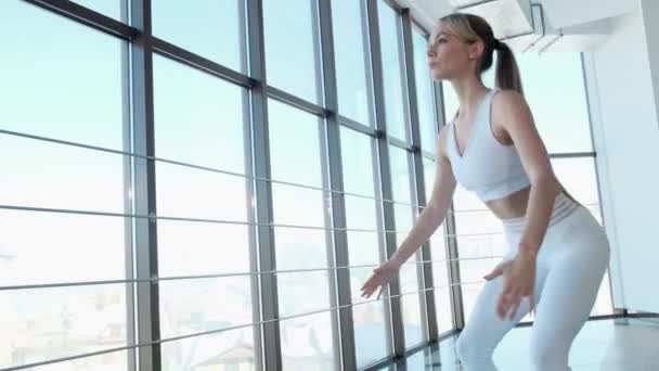 A young athlete does squats near a large bright window, a fitness trainer — Stock Video
