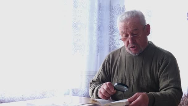 Hombre mayor con gafas lee un libro junto a la ventana en casa. — Vídeos de Stock