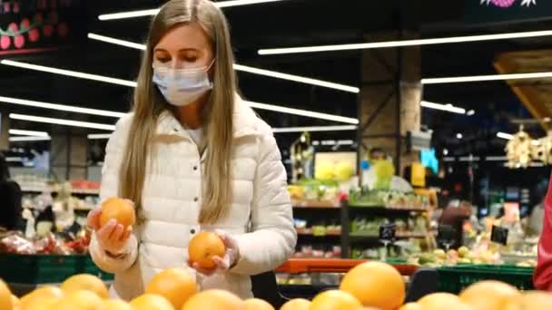 Μια γυναίκα με προστατευτική μάσκα στο σούπερ μάρκετ αγοράζει πορτοκάλια. Φρούτα στο σούπερ μάρκετ. — Αρχείο Βίντεο