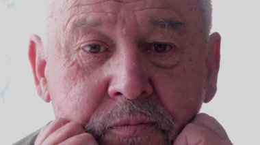 Gri saçlı yaşlı bir adam, elleri yüzüyle düşünceli bir şekilde oturur.