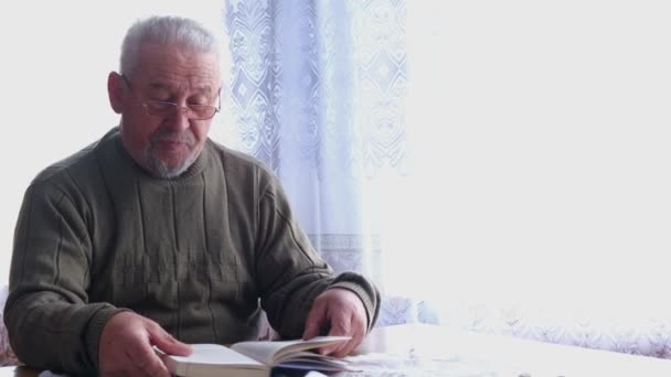 Een gepensioneerde met een bril die een boek leest terwijl hij op een stoel zit in het Huis — Stockvideo