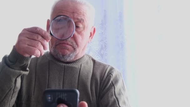 Abuelo lee las noticias en un teléfono inteligente con una lupa — Vídeo de stock