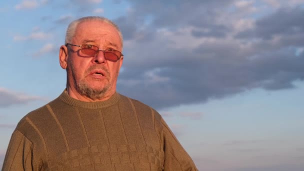 Um homem velho em uma camisola cinza e óculos de sol contra um céu azul. — Vídeo de Stock