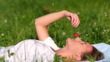 Genç bir kız yeşil çimlere uzanıyor ve olgun kırmızı kirazı tadıyor..