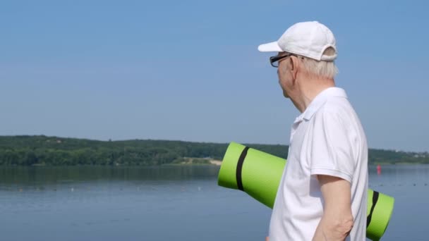 Farfar gick i pension i morgon stadspark, håller en yogamatta, tittar på sjön. — Stockvideo