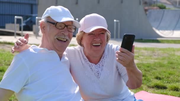 Os aposentados sorridentes e felizes usam um smartphone enquanto estão sentados no parque na grama. — Vídeo de Stock