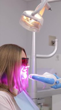 Dikey video. Bir hasta modern, gelişmiş bir diş kliniğinde LED ışığı kullanılarak diş beyazlatma prosedüründen geçiyor.
