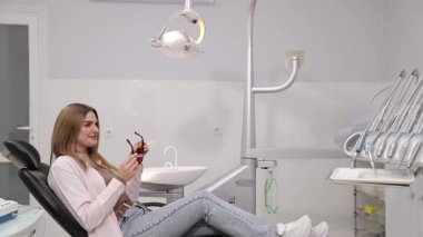 Genç bir kadın, dişçi koltuğunda rahat bir şekilde oturur ve klinik kontrolü sırasında şık bir güneş gözlüğü takar.