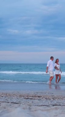 Bir çift deniz kıyısında yavaşça yürüyor, sohbet ediyor ve gülümsüyor birlikte sakin ve huzurlu bir sahil anın tadını çıkarıyor..