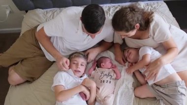 İki çocuklu ebeveynler, yeni doğan bebeklerine gülümseyerek yatakta yan yana yatıyorlar. Güzel bir bağ, şefkat ve sıcaklığın doğal ışıkla yakalandığı bir an..