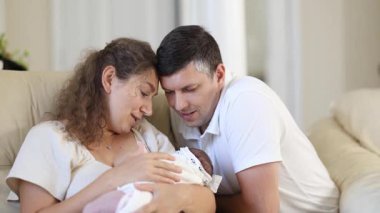 Gülümseyen anne ve baba, güneşli bir oturma odasındaki kanepede otururken yeni doğan bebeklerine sevgiyle bakıyor. Duygusal aile bağları neşe, şefkat ve bağlantı gösteriyor..