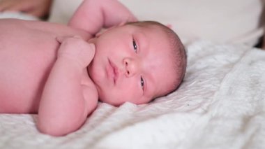 Sevimli yeni doğmuş bebek beyaz bir yatakta uzanıyor, sakince dinleniyor ve merakla kameraya bakıyor. Yumuşak ışıklar ve yumuşak tonlar masumiyeti, şefkati ve erken yaşam güzelliğini vurgular..