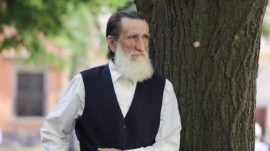 Gri saçlı yaşlı bir adam güneşli bir günde modern şehir sokaklarında tek başına yürüyor. Sakin kentsel atmosfer emeklilikte günlük hayatı, yaşı ve özgürlüğü gösteriyor. Yaşam tarzı, seyahat ve son sınıf konsept projeleri için mükemmel.