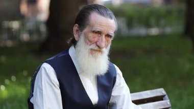Uzun beyaz sakallı ve arkaya taranmış saçları olan yaşlı bir adam, siyah yelek ve beyaz gömlek giymiş bir bankta oturur, düşünceli bir şekilde kameraya bakar, sakin ve bilgeliği yakalar.