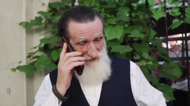 Beyaz sakallı ve bıyıklı yaşlı bir adam telefonuyla konuşuyor. Yoğun yeşil yaprakların yanında duruyor. Açık havada demir parmaklıklar var..