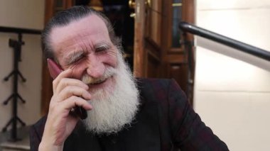 Beyaz sakallı kıdemli bir adam içeride telefonla konuşuyor. Çağrı sırasında gülümsüyor ve el kol hareketi yapıyor..