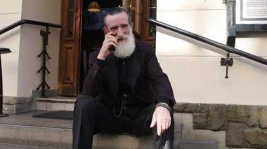 Beyaz sakallı yaşlı bir adam dışarıda merdivenlerde otururken telefonla konuşuyor. Çağrı sırasında doğal bir şekilde gülümsüyor ve el kol hareketi yapıyor..