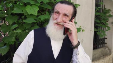 Uzun beyaz sakallı yaşlı adam şehir kaldırımında yemyeşil çalıların yanında duruyor, siyah yelek ve beyaz gömlek giyerken cep telefonuyla konuşuyor..