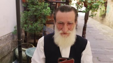 Uzun beyaz sakallı yaşlı bir adam akıllı telefonunu kullanırken açık bir kafede ya da sokak bankında oturur. Sahne, şehir ortamında teknoloji, iletişim ve modern yaşam tarzlarını kucaklayan yaşlıları yakalıyor..