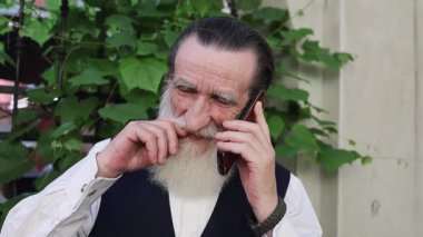 Uzun beyaz sakallı yaşlı adam sık yeşilliklerin yanında duruyor ve bir ara sokakta bir pencere, cep telefonuyla konuşuyor ve günlük yaşlıları resmediyor..