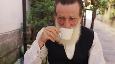 Uzun beyaz sakallı yaşlı bir adam dışarıda bir kafede ya da terasta otururken beyaz bardaktan yudumluyor. Huzurlu bir an, rahatlama, rutin ve sıcak bir içeceğin tadını çıkarma.. 