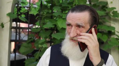 Uzun beyaz sakallı yaşlı bir adam yemyeşil sarmaşıkların ve demir bir kapının yanında duruyor ve cep telefonuyla konuşuyor. Koyu renk bir yelek ve beyaz bir gömlek giyiyordu. Bağlantı ve günlük yaşamı aktarıyordu..