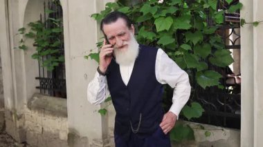 Uzun beyaz sakallı yaşlı adam, siyah yelek ve beyaz gömlek giyerken cep telefonuyla konuşan, sarmaşık kaplı taş bir duvara yaslanıyor..