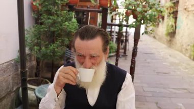 Uzun beyaz sakallı yaşlı adam bir fincan kahveyle açık bir kafede oturuyor, akıllı telefonunu kullanıyor ve huzurlu bir anın tadını çıkarıyor..