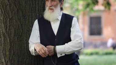 Siyah yelekli, uzun beyaz sakallı yaşlı adam şehir parkında büyük bir ağaç gövdesinin yanında duruyor. Sakince etrafına bakıyor ve huzurlu bir açık hava anının tadını çıkarıyor..