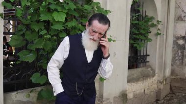 Uzun beyaz sakallı, koyu renk yelekli yaşlı bir adam yeşil asmalarla kaplı bir bina boyunca yürüyor ve cep telefonuyla konuşuyor..