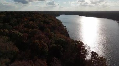 Hava görüntüleri, renkli düşen ağaçlarla çevrili huzurlu bir göl gösteriyor. Sakin su yüzeyine yansıyan ılık ışık, sakin bir sonbahar havası yaratır..