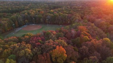 İHA görüntüleri, parlak kırmızı, turuncu ve sarı ağaçlarla çevrili küçük bir beyzbol sahası gösteriyor. Sonbahar mevsiminde huzurlu Amerikan kırsal arazisi.