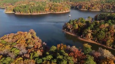 İnsansız hava aracı görüntüleri, sarı, kırmızı ve yeşil yapraklı ağaçlarla çevrili huzurlu mavi bir göl gösteriyor. Güzel yansımalar ve sıcak sonbahar renkleri.