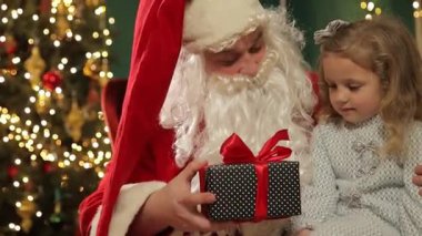 Noel Baba, parıldayan bir Noel ağacının yanında genç bir kıza nazikçe bir hediye veriyor ve sıcak bir tatil sahnesi yaratıyor..