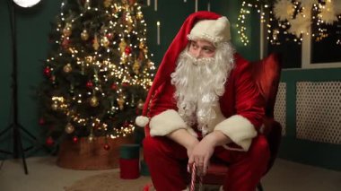 Noel Baba, hediyeler ve sıcak şenlik süslemeleriyle dolu parıldayan bir Noel ağacının yanında sessizce oturur..