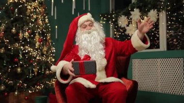 Noel Baba ışıklarla ve hediyelerle çevrili Noel ağacının yanında rahat bir sandalyede otururken mutlu bir şekilde el sallıyor..