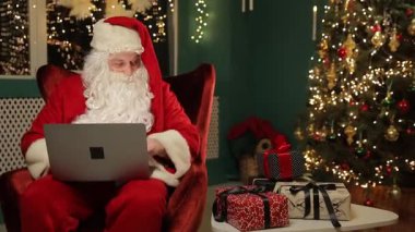 Noel Baba hediye listesini hediye ve ışıklarla dolu parlayan bir Noel ağacının yanındaki dizüstü bilgisayarda dikkatlice gözden geçiriyor..